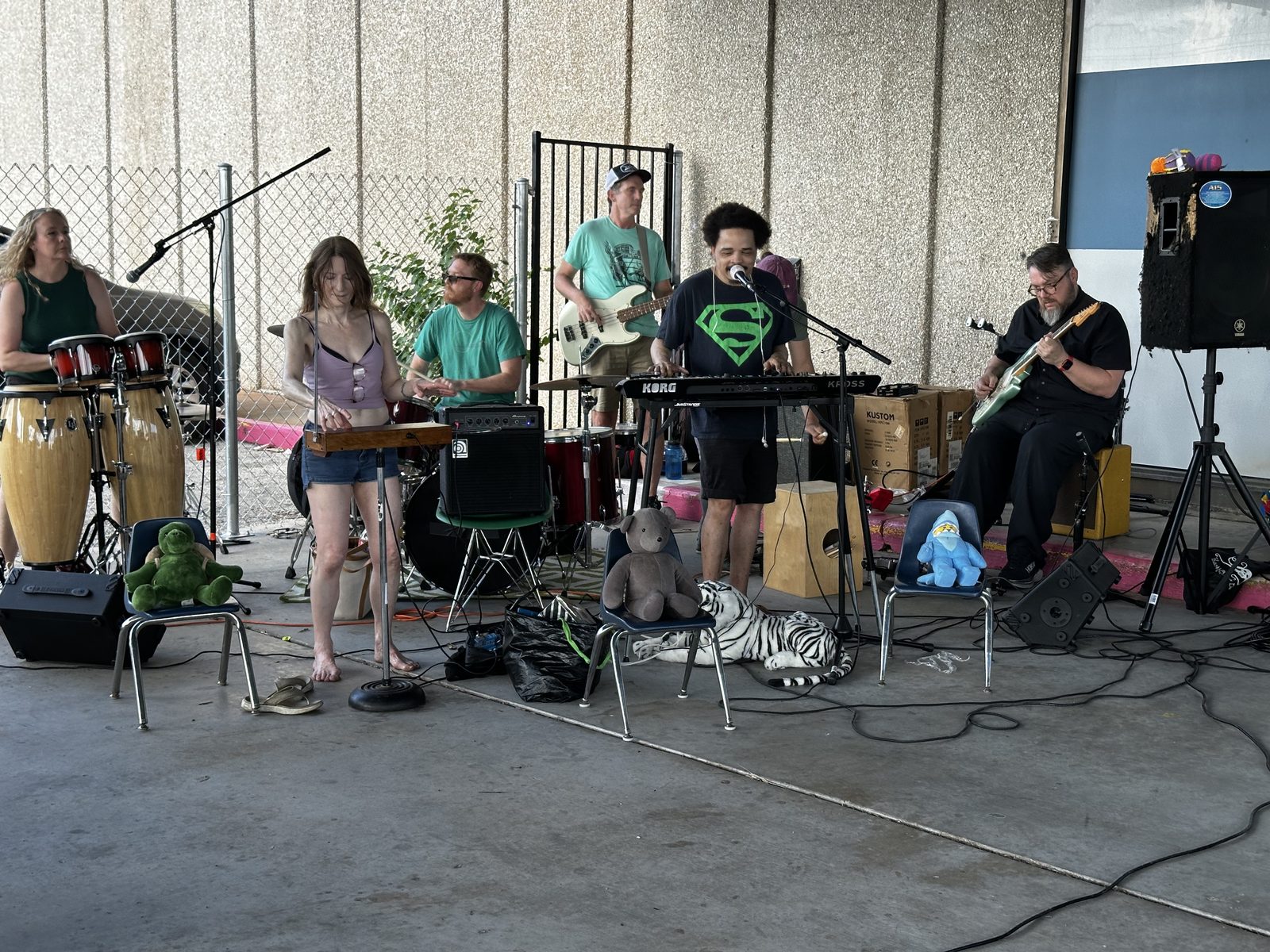 The band Ouiness playing under a carport, seven of them, with stuffed animals watching from child-sized chairs.