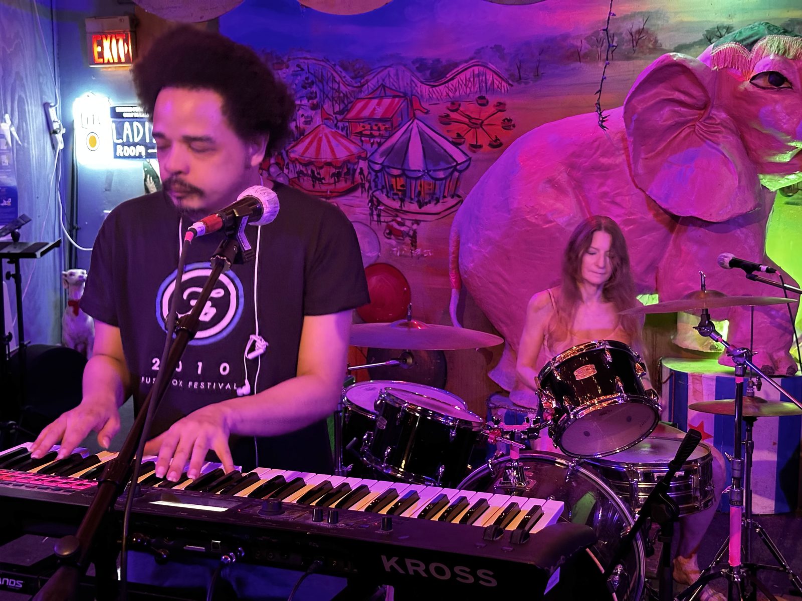 T.J. Wade on keys and Anne on drums, the band Line of Fire playing an Austin bar under a carnival mural and a giant pink elephant.