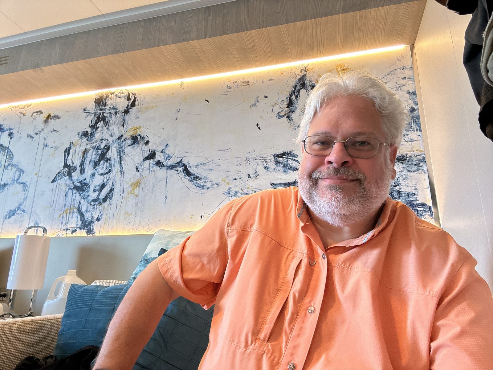 Ernie in an orange shirt in his stateroom at Antigua, a large abstract painting behind him.